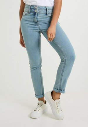 Pantalones vaqueros de tiro alto en un azul claro con un corte ajustado, cierre de dos botones y puños enrollados, combinados con zapatillas blancas con detalles en dorado.