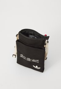 Svart kanvasveske med glidelås på toppen, beige stropp og frontlommene. Har hvit brodert tekst og et logoaccent på bunnen.