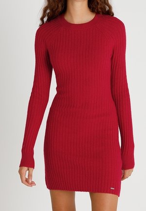 Rode ribgebreide jurk met lange mouwen, voorzien van een ronde hals en een nauwsluitende pasvorm. Klein logo detail aan de zoom, zachte textuur.