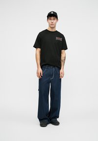 T-shirt en coton noir avec un logo graphique, associé à un pantalon large à rayures bleu marine. Le modèle porte une casquette noire et des chaussures sombres.