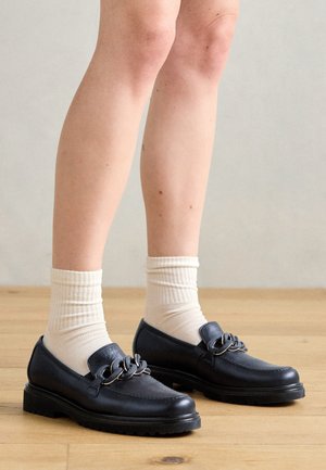 Jambes portant des chaussettes blanches et des mocassins en cuir noir avec un grand détail chaîne, debout sur un sol en bois contre un mur uni.