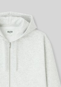 Haut à capuche zippé gris clair en tissu doux. Doté d'une capuche, d'une fermeture éclair avant et d'une texture subtile. Marque visible sur l'étiquette intérieure.