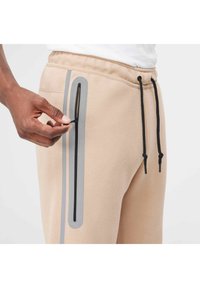 Pantaloni jogger beige con una tasca laterale nera con zip e strisce grigie di contrasto. Vita elastica con lacci neri e tessuto testurizzato.