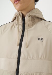 Lys beige jakke med hætte, der har en sort lynlås, elastiske detaljer og et lille "P.E. Nation" logo på brystet. Glat tekstur.