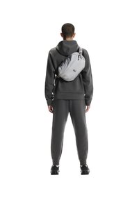 Graue Umhängetasche mit Reißverschluss, verstellbarem Riemen und Logodetail, getragen über einem passenden grauen Hoodie und einer Jogginghose.