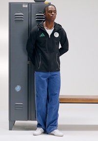 Black zip-up jacket with green accents, blue denim jeans, and white sneakers, standing beside a gray locker and wooden bench.