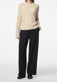 Kvinna som bär en beige stickad tröja, vida svarta byxor och svarta glänsande skor, står med ena handen i fickan.