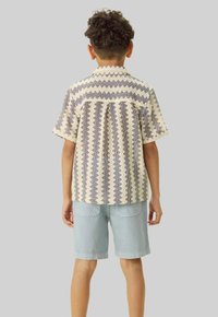 Chemise à manches courtes, à motif, avec une base crème et un motif en zigzag bleu, associée à un short en denim clair, vue de dos.