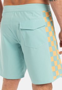 Tyrkysové plavkové šortky s bočnou vreckom, so žlto-modrým šachovnicovým vzorom na boku, vyrobené z mäkkej tkaniny a s prešitým lemom.