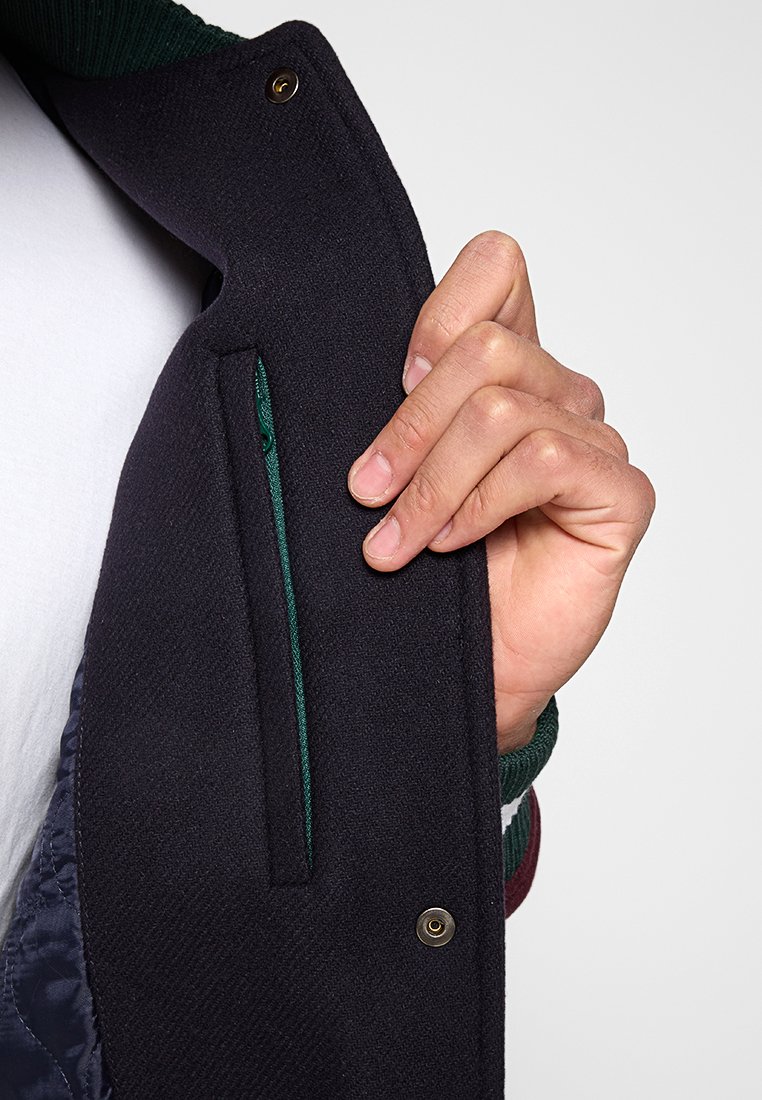 Veste marine en tissu texturé, dotée d'une poche zippée doublée de vert. Le col et les poignets ont un accent en tricot côtelé.