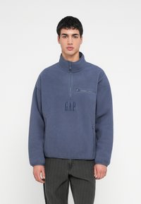 Młody mężczyzna ubrany w niebieską polarową bluzę z półzamkiem, z kieszenią na piersi na zamek i logo "GAP", w połączeniu z ciemnoszarymi jeansami.