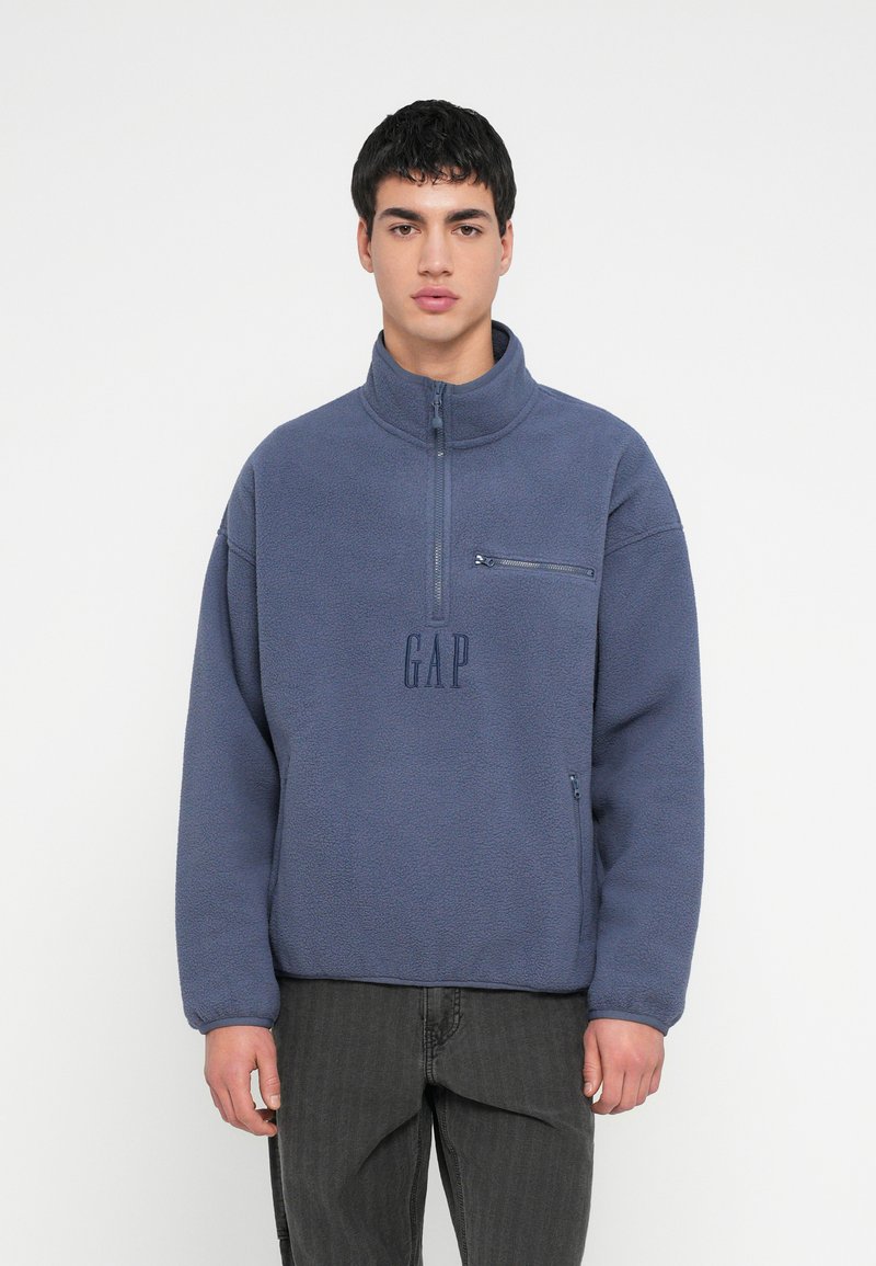 Młody mężczyzna ubrany w niebieską polarową bluzę z półzamkiem, z kieszenią na piersi na zamek i logo "GAP", w połączeniu z ciemnoszarymi jeansami.