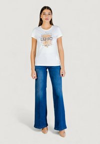 Camiseta blanca de manga corta con un gráfico colorido de flores y texto en el centro. Combinada con jeans de mezclilla azul acampanados y zapatos beige.