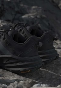 Paire de chaussures de randonnée noires avec semelles épaisses sur un sol rocheux, mettant en valeur l'adhérence texturée et les languettes de traction aux talons.