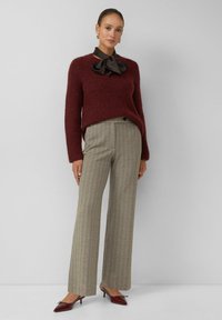 Femme debout portant un pull bordeaux, un nœud de cou en satin noir, un pantalon beige à jambe large et des talons aiguilles bordeaux sur un fond uni.