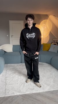 Zwarte rits-hoodie met grafisch logo, gecombineerd met zwarte sweatpants met een klein ontwerp. Lichtgekleurde sneakers op een zachte ondergrond.