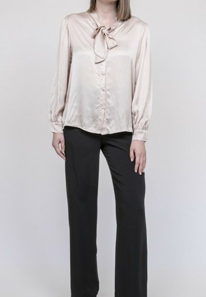 Femme portant un chemisier en satin rose clair à col lavallière et un pantalon noir large à jambes longues, debout devant un fond gris uni.