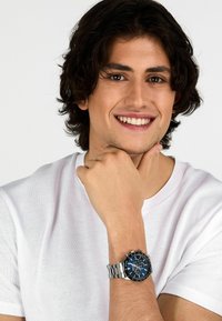 Orologio in acciaio inossidabile argentato con quadrante blu, dotato di funzionalità cronografo e bracciale in metallo, indossato al polso di un modello con una t-shirt bianca.