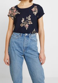 Námořnický květinový top s krátkými rukávy, se zaobleným výstřihem a texturovanou tkaninou. Top má vzory světle růžových a béžových květin. Nosený s modrými džíny vysokého pasu.