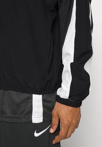 Man wearing black and white sportswear with long sleeve jacket and black pants featuring a white Nike logo and white vertical stripes.