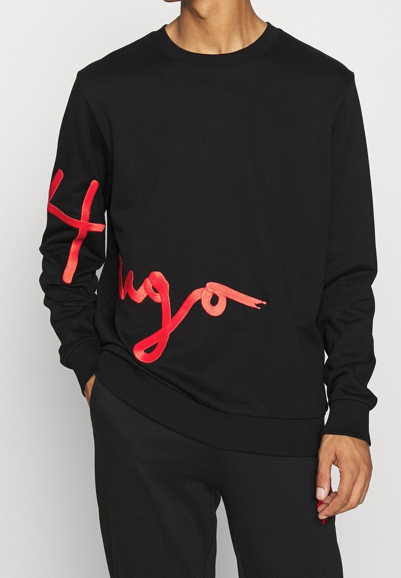 Homme portant un sweat-shirt noir avec des lettres cursives rouges sur le côté gauche et sur la manche droite, debout avec une main dans la poche.