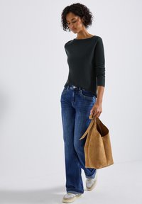Maglione fitted verde scuro, jeans a gamba larga blu e borsa a tracolla in suede beige. Sneakers chunky crema con dettagli marrone chiaro. Capelli ricci, sfondo neutro.