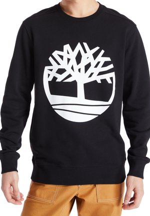 Homme portant un sweat-shirt noir avec un grand logo d'arbre stylisé blanc centré sur la poitrine, associé à un pantalon marron clair.