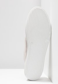 Zapato con una parte superior de ante color beige claro y suela de goma blanca. La suela presenta un patrón de zigzag para mayor tracción. Tamaño marcado en un lado.