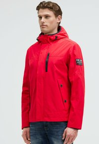 Homme portant une veste à capuche rouge vif avec une fermeture éclair et des boutons noirs, se tenant devant un fond clair.