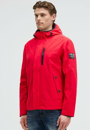 Man wearing bright red hooded jacket with black zipper and buttons, standing against plain light background.