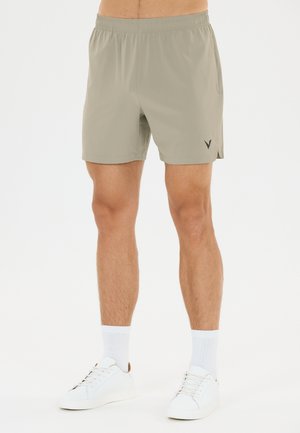 Mand iført beige atletikshorts med et lille logo, hvide sokker og hvide sneakers mod en ensfarvet hvid baggrund.