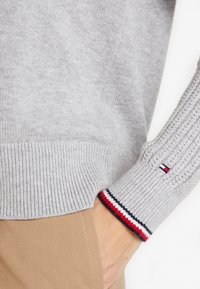 Light gray knit sweater cuff with red, white, and navy stripes above beige pants, showing a hand partially in pocket.