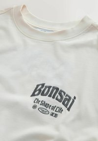 Roomkleurig katoenen T-shirt met een geribde ronde hals en geborduurde zwarte tekst: "Bonsai - The Shape of Life" met grafische accenten.