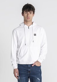 Biela mikina na zips vyrobená z mäkkej tkaniny, s prednou kapsou, šnúrok a malým čiernym logom na ľavom hrudníku. Nosí sa s modrými džínsami.