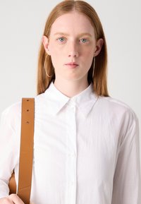 White button-up shirt with a collar and front placket, featuring a textured, slightly wrinkled fabric. Brown shoulder strap visible.