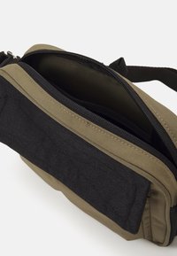 Midjeväskan i olivgrön och svart tyg, med ett huvudfack som stängs med dragkedja och en texturerad svart panel fram.