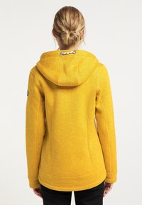 Gelbe Kapuzenjacke mit strukturierter Oberfläche, ausgestattet mit einer Kapuze, Seitennähten und einem sichtbaren Logo-Patch am Ärmel.