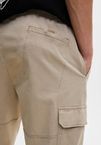 Beige Cargo-Shorts mit hinteren und seitlichen Klappentaschen, getragen von einer Person, die eine Hand in der Seitentasche hat, mit detaillierten Nähten und Stoffstruktur.