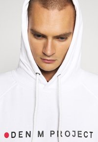 Balta hoodie su raišteliu, ant krūtinės esančiu užrašu "DENIM PROJECT" juodu tekstu. Minkšta medžiagos tekstūra.