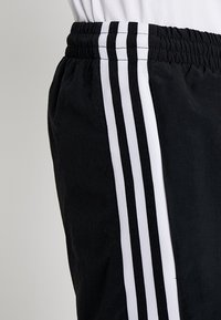 Svarta träningsbyxor med ett elastiskt midjeband, som har tre vertikala vita ränder längs sidan. Materialet verkar glatt och lätt.