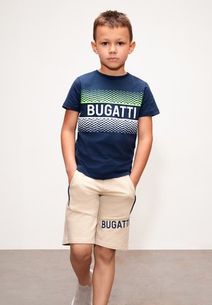 T-shirt blu navy con un motivo verde e bianco con la scritta "BUGATTI", abbinata a pantaloni beige con il logo "BUGATTI" sulla gamba.