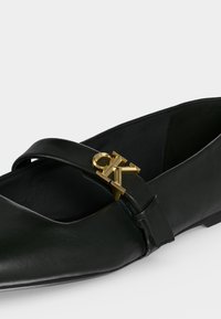 Chaussure plate en cuir noir avec un bout pointu et une boucle métallique dorée avec le logo CK sur la bride du dessus.