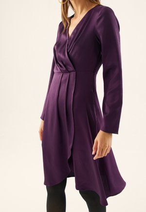 Kobieta w długiej sukience kopertowej o głębokim purpurowym kolorze, z plisami i asymetrycznym dołem, w połączeniu z czarnymi rajstopami.