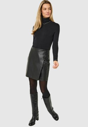Vrouw in een zwarte coltrui, zwarte leren wikkelrok, zwarte panty en kniehoge zwarte laarzen, staand tegen een effen lichte achtergrond.