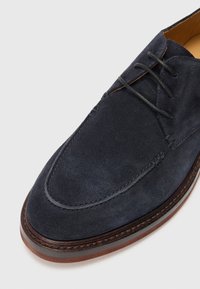 Granatowae zamszowe buty z okrągłym czubkiem, z sznurówkami, kontrastowym przeszyciem i brązową gumową podeszwą z lekkim obcasem.