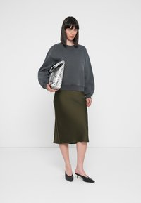 Gray, oversized sweatshirt with round neck, paired with a dark green satin midi skirt. Holding a textured silver snake-print clutch. Black mules.