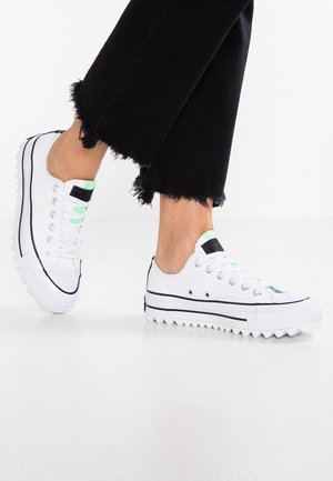 White low-top sneakers with black trim, thick jagged soles, and green accents worn with black frayed-hem pants, on a white background.