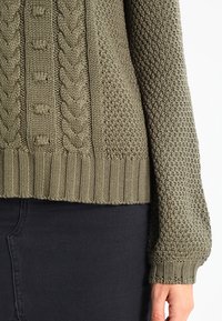 Pull tricoté vert olive avec un motif texturé, un design en torsades sur le devant, et des poignets et une bordure côtelés.