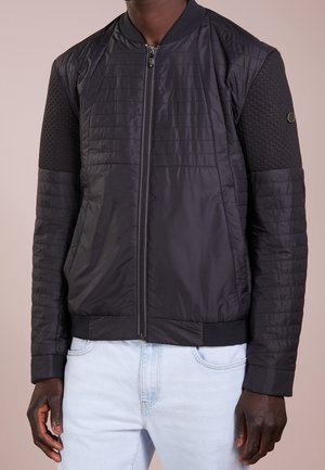 Mężczyzna ubrany w czarną, pikowaną kurtkę bomberkę z fakturowanymi rękawami i jasnoniebieskie dżinsy, stojący na tle o neutralnym kolorze.