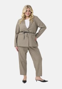 Beige tailored blazer with a tie belt, paired with matching trousers. Blazer features a notched collar, and both pieces have a smooth texture.